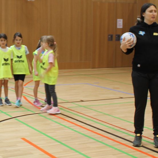 Eine Frau, die einen Ball hält, steht in einer Turnhalle mit vier Mädchen in gelben Trikots. Die Mädchen stehen in einer Reihe und schauen die Frau an.