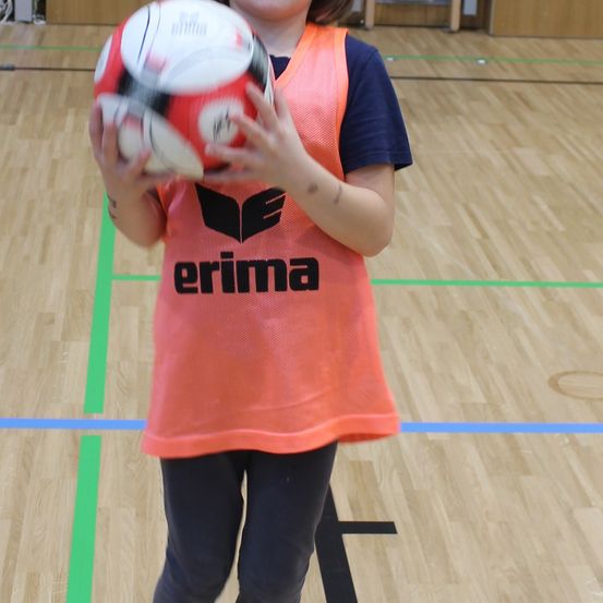 Ein junges Kind in einem orangefarbenen Erima-Trikot hält einen Fußball in einer Turnhalle. Der Boden hat blaue und grüne Markierungen.