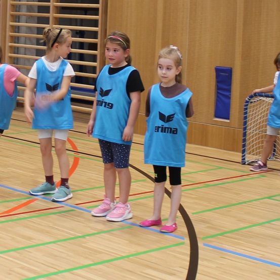 Eine Gruppe junger Mädchen in passenden blauen Uniformen steht auf einem Spielfeld, eine hält einen Stock und eine andere steht neben einem Tor. Das Spielfeld ist mit bunten Linien markiert.