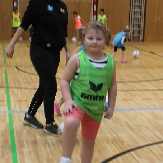 Eine Frau in Schwarz und ein junges Mädchen in Grün spielen Fußball auf einem Hallenplatz. Hinter ihnen laufen Kinder und spielen mit Fußballbällen. Der Platz hat Holzboden und Markierungen.
