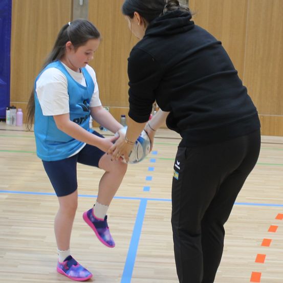 Ein Mädchen übt mit einem Ball in einer Turnhalle. Eine Frau leitet sie. Sie stehen auf einem Holzboden mit blauen Markierungen.