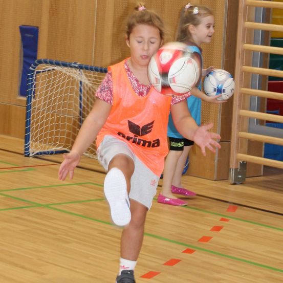 Zwei junge Mädchen spielen Fußball in einer Turnhalle, ein Mädchen tritt einen Ball, während das andere einen Ball hält.
