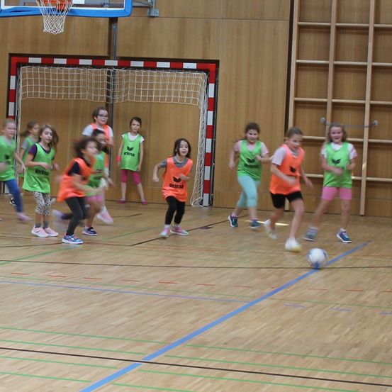Eine Gruppe von Kindern spielt Fußball auf einem Hallenplatz. Sie tragen grüne und orange Trikots. Der Platz hat blaue Markierungen und ein Basketballtor im Hintergrund.