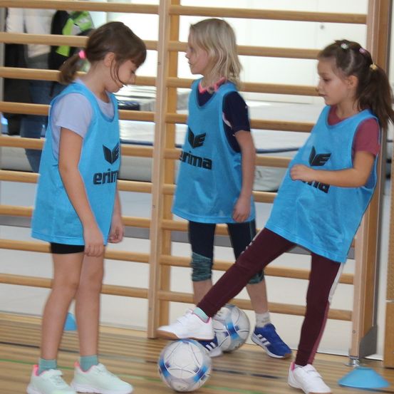 Drei junge Mädchen in blauen Trikots spielen drinnen Fußball, wobei ein Mädchen den Ball tritt und die anderen zusehen. Im Hintergrund befinden sich Holzstangen und Matten.
