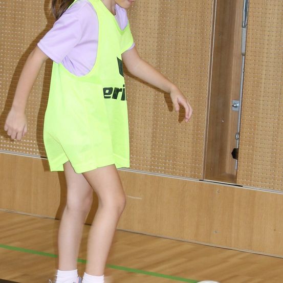 Ein junges Mädchen in einem gelben Westchen steht auf einem Spielfeld mit ausgestreckten Händen. Die Wand hinter ihr hat eine hölzerne Textur.