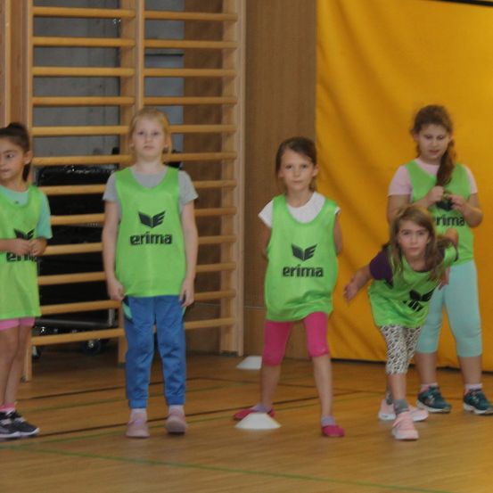 Fünf junge Mädchen in grünen Trikots mit dem Wort 'erima' stehen auf einem Holzboden, möglicherweise in einer Turnhalle.
