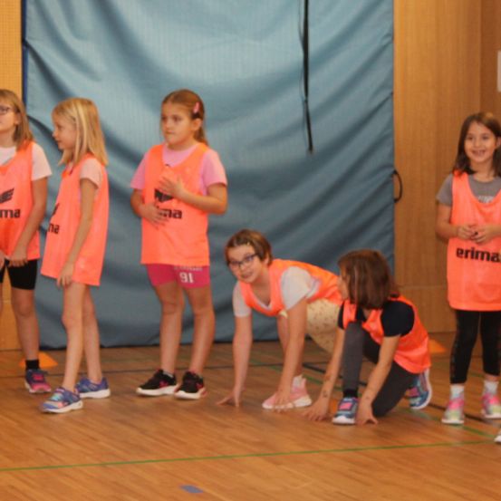 Eine Gruppe junger Mädchen steht in einer Turnhalle in orangefarbenen Westen, eine Mädchen hockt, während die anderen stehen und zusehen.