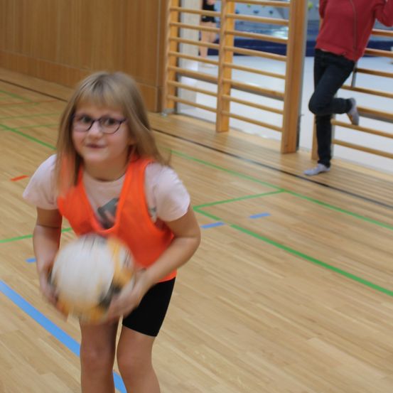 Ein Mädchen in einem orangefarbenen Weste hält einen Fußball in einer Turnhalle mit Holzböden und grünen Markierungen. Eine Person in Rot und Schwarz ist im Hintergrund verschwommen.