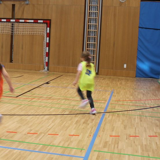 Ein verschwommenes Bild von zwei Kindern, die auf einem Hallenplatz Fußball spielen, mit einem Tor und einer Leiter im Hintergrund.