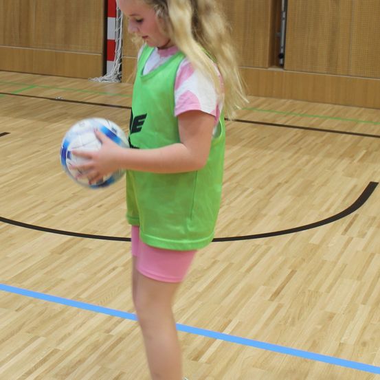 Ein Mädchen in einem grünen Weste und rosa Shorts hält einen Fußball in einer Turnhalle mit Holzboden und grünen Linien.