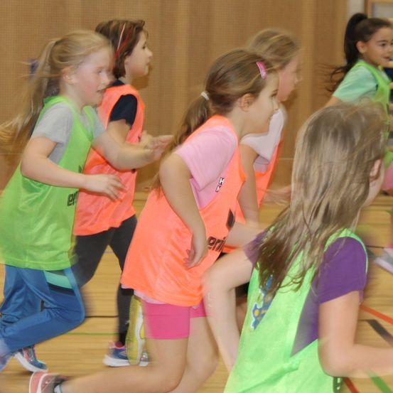 Eine Gruppe junger Mädchen in Sportkleidung läuft drinnen. Sie tragen unterschiedlich farbige Westen. Ihre Haare sind zurückgebunden, und sie scheinen an einem Spiel beteiligt zu sein.
