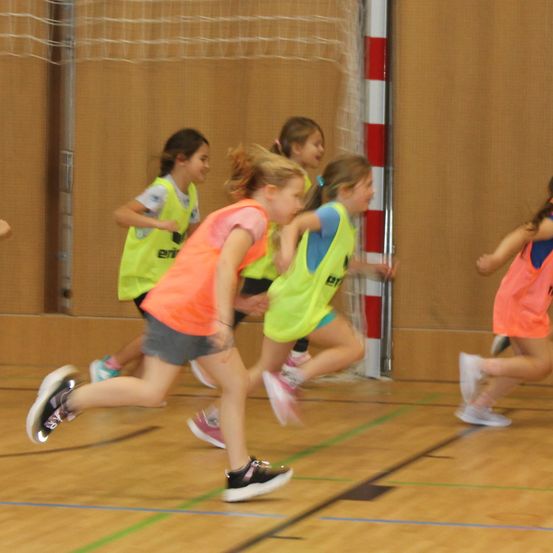 Eine Gruppe junger Mädchen läuft in einer Turnhalle und trägt helle gelbe und orangefarbene Westen. Sie sind verschwommen, was auf schnelle Bewegung hinweist.