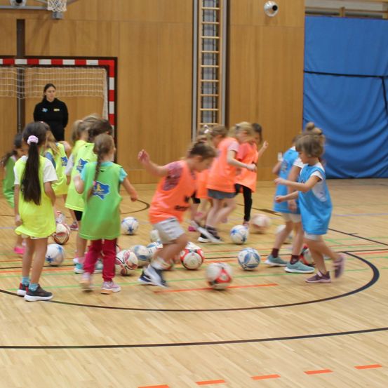Eine Gruppe junger Mädchen in passenden Trikots spielt mit Fußballbällen auf einem Hallenplatz. Eine Frau in einem schwarzen Shirt beobachtet von der Seite.