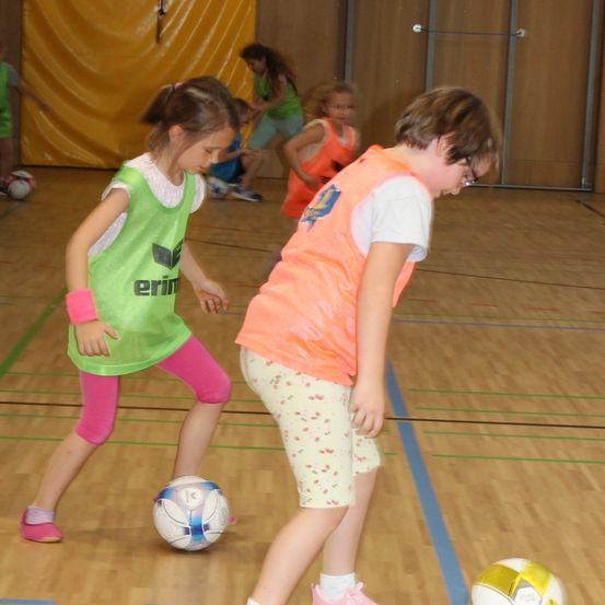 Kinder spielen Fußball auf einem Indoor-Spielfeld. Ein Mädchen in einem grünen Shirt hält einen Fußball. Ein anderes Kind in einem orangefarbenen Shirt beobachtet den Ball.