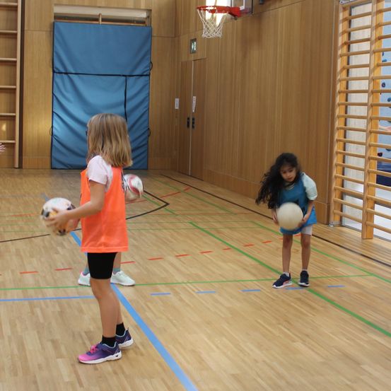 Zwei junge Mädchen spielen mit Bällen in einer Turnhalle. Ein Mädchen trägt eine orange Weste und das andere ein blaues Shirt. Der Boden ist mit Linien markiert.
