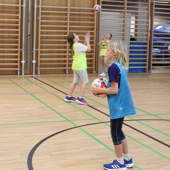 Drei Mädchen spielen Ball in einer Turnhalle mit Holzboden und grünen Markierungen. Ein Mädchen in einem gelben Shirt streckt die Hand nach dem Ball aus, während ein anderes in einem blauen Shirt ihn hält. Ein Mädchen in einem gelben Shirt steht hinter ihnen.