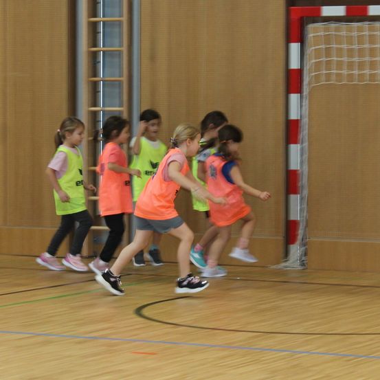 Eine Gruppe junger Kinder in neonorangenen Westen rennt auf einem Hallenplatz in der Nähe eines Tornetzes.