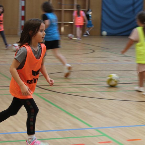 Ein junges Mädchen in einem orangefarbenen Trikot läuft auf einem Hallenspielfeld. Hinter ihr spielen mehrere andere Kinder Fußball. Das Spielfeld hat blaue und grüne Linien.