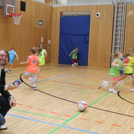 Eine Gruppe von Kindern spielt Fußball in einer Turnhalle. Ein Mann beobachtet sie und hält eine Flasche. Ein Basketballkorb ist an der Wand.