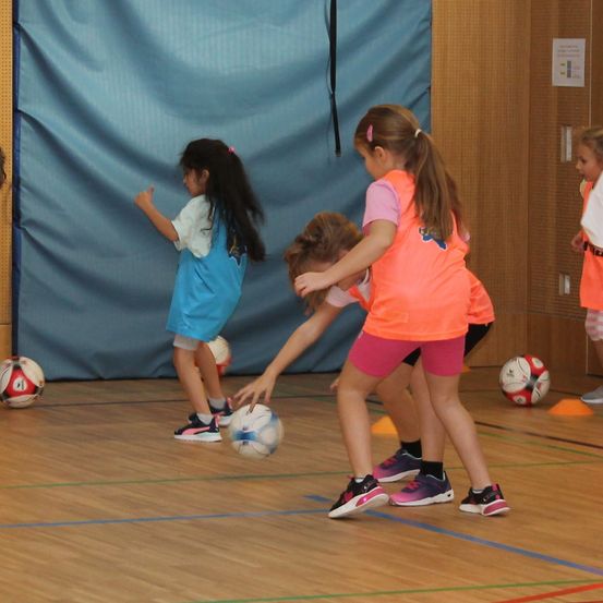 Drei Mädchen in orangefarbenen und blauen Trikots spielen mit Fußballbällen auf einem Spielfeld. Ein Mädchen tritt einen Ball, während ein anderes sich beugt, um ihn zu fangen. Dahinter ist eine blaue Wand und einige Verkehrskegel zu sehen.