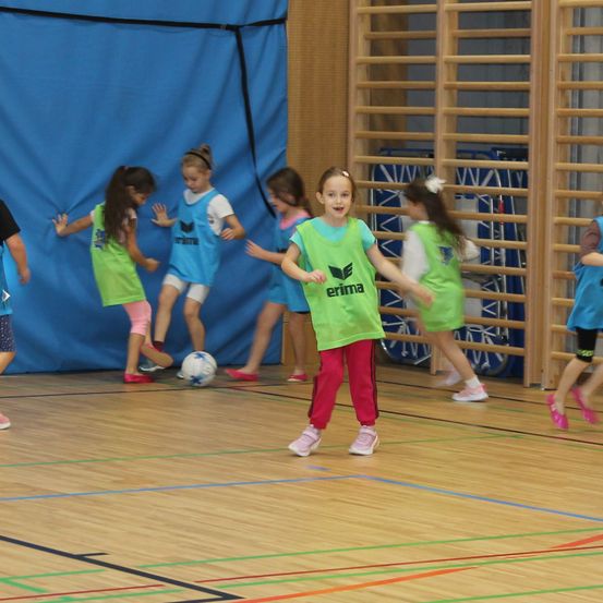 Mehrere junge Mädchen spielen Fußball in einer Turnhalle. Sie tragen passende grüne Trikots mit 'erima' auf ihren Jerseys. Ein Mädchen tritt einen Fußball, während andere zusehen.