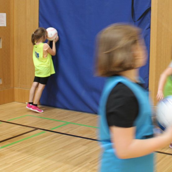 Ein junges Mädchen übt das Werfen eines Balls in einer Turnhalle. Sie hält den Ball gegen eine blaue Wand. Ein weiteres Kind ist im Vordergrund verschwommen.