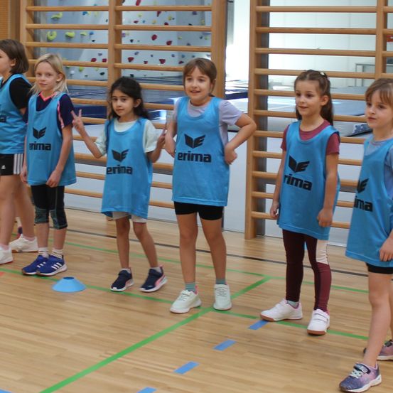 Mehrere junge Mädchen stehen auf einem Turnhallenboden und tragen identische blaue Trikots mit dem Wort 'erima' darauf. Sie stehen mit blauen Hütchen zwischen ihnen aufgereiht, und im Hintergrund befindet sich eine Kletterwand.