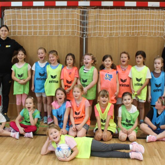 Eine Gruppe junger Mädchen in leuchtenden Trikots posiert für ein Foto vor einem Fußballtor, mit einer Frau dahinter.