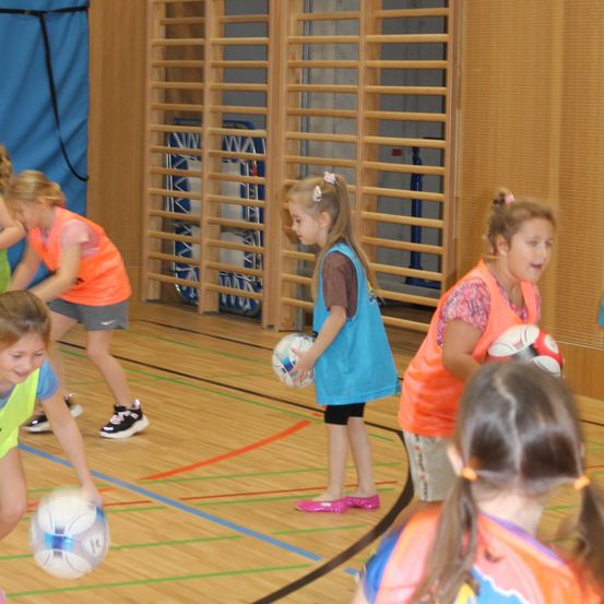 Eine Gruppe junger Mädchen spielt mit Fußballbällen in einer Turnhalle. Sie tragen Sporttrikots und Turnschuhe. Eines der Mädchen hält einen Ball, und ein anderes schaut sie an.