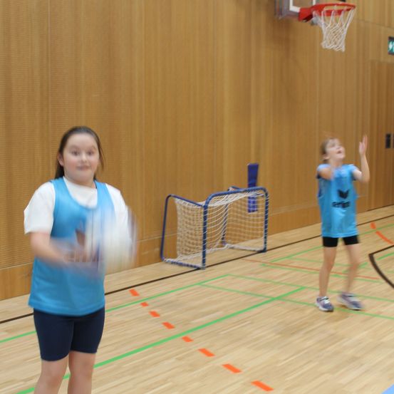 Zwei Mädchen spielen Basketball in der Halle. Ein Mädchen wirft den Ball in Richtung Korb, während das andere ihn fängt. Die Halle hat Holzboden und einen Basketballkorb.