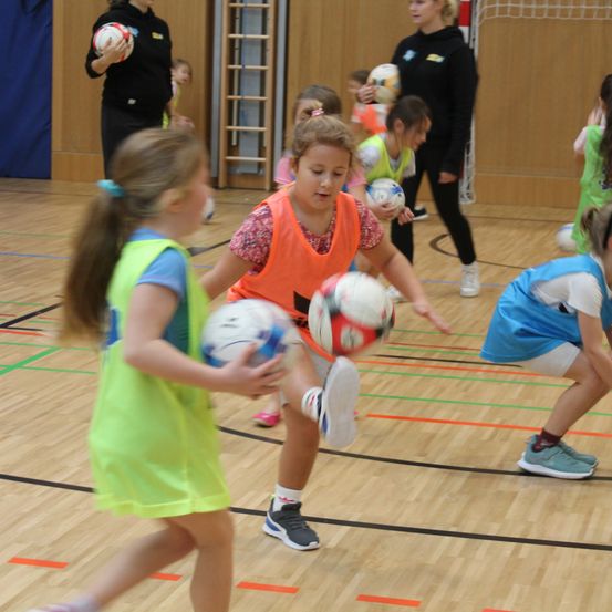 Eine Gruppe junger Mädchen spielt in einer Turnhalle ein Ballspiel. Einige tragen Westen und Turnschuhe.