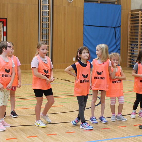 Eine Gruppe junger Mädchen in orangefarbenen Trikots steht in einer Turnhalle, wahrscheinlich in Vorbereitung auf ein Spiel. Sie tragen alle Turnschuhe und blicken nach vorne.