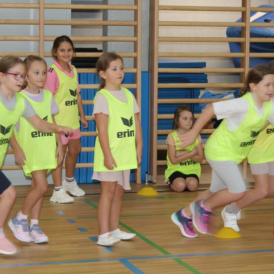 Eine Gruppe junger Mädchen in neonfarbenen Westen spielt ein Spiel auf einem Sportplatz. Einige stehen, andere hocken, mit gelben Kegeln verstreut.