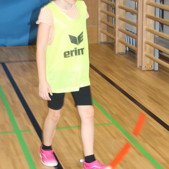 Ein junges Mädchen in einem gelben Weste mit 'erim' darauf steht auf einem Holzboden mit grünen und orangefarbenen Linien.