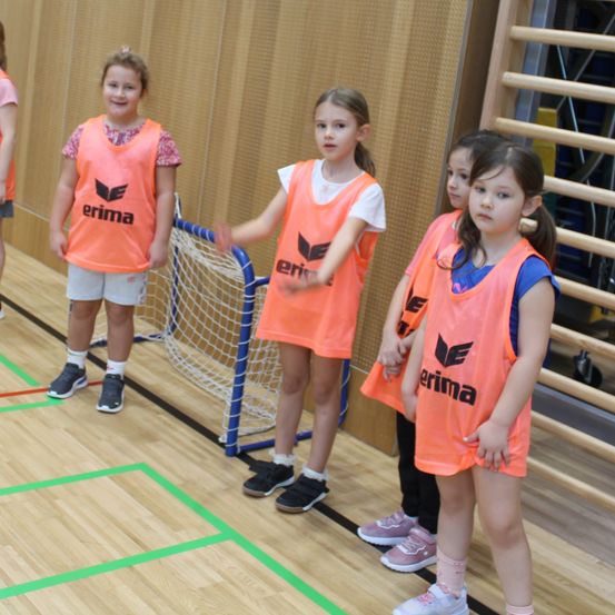 Fünf Mädchen in orangefarbenen Trikots stehen auf einem Spielfeld, wahrscheinlich in einer Sporthalle. Sie tragen Turnschuhe und einige haben Socken an. Ein Mädchen vorne gestikuliert. Ein blaues Tor steht in der Nähe.