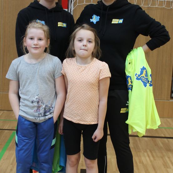 Drei junge Mädchen stehen lächelnd zusammen in einer Turnhalle. Die Mädchen tragen verschiedenfarbige Shirts und Hosen. Hinter ihnen stehen zwei Erwachsene und halten gelbe T-Shirts mit Logos. Die Turnhalle hat Holzböden und eine Wand mit einem Netz.