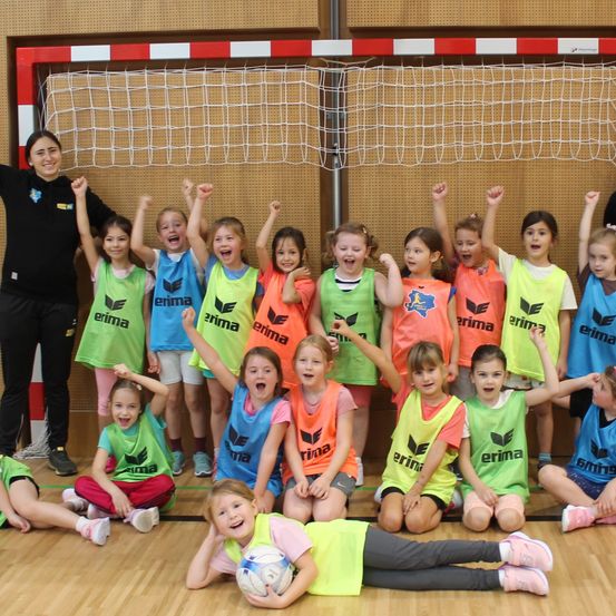 Eine Gruppe junger Mädchen in Sportuniformen posiert für ein Foto in einer Turnhalle. Eine Frau steht hinter ihnen in der Nähe eines Tornetzes.