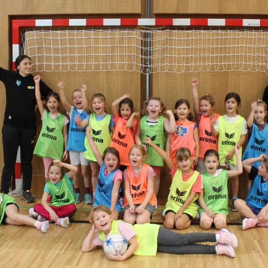 Eine Gruppe junger Mädchen in leuchtenden Trikots posiert für ein Foto in einer Turnhalle. Der Trainer steht vor einem Netz.