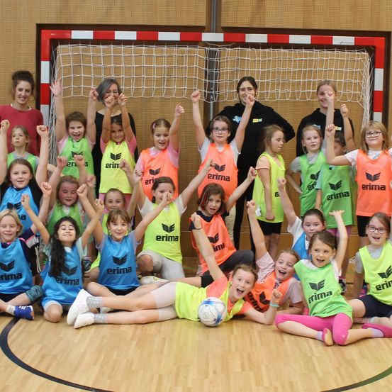 Eine Gruppe junger Mädchen und ihre Trainer feiern gemeinsam vor einem Handballtor. Sie tragen leuchtende Trikots mit dem Wort 'erima' darauf.