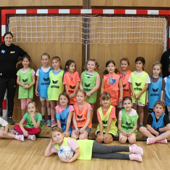 Eine Gruppe junger Mädchen in Sporttrikots posiert für ein Foto vor einem Tor. Eine Frau steht hinter ihnen.