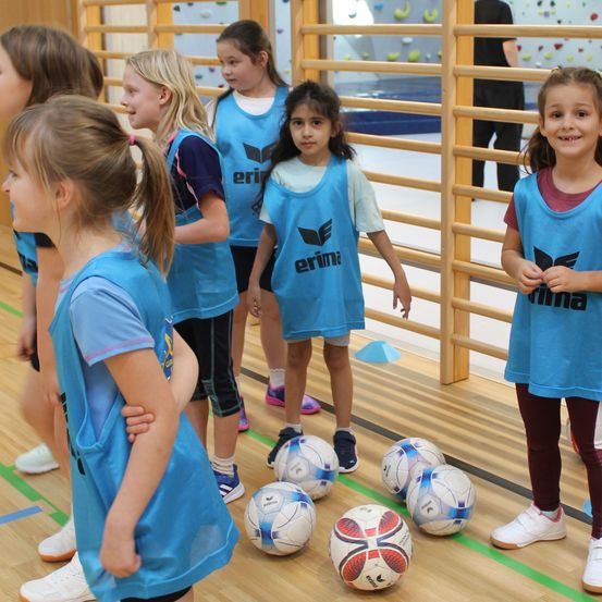 Eine Gruppe junger Mädchen in blauen Uniformen steht in einer Sporthalle und blickt auf eine Reihe von Fußballbällen auf dem Boden. Im Hintergrund sind Klettergeräte zu sehen.