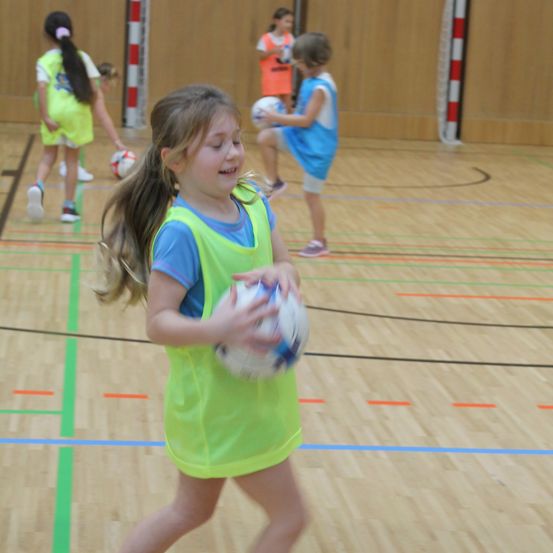 Eine Gruppe junger Mädchen spielt eine Partie Fußball in der Halle. Das Mädchen vorne läuft mit einem Fußball.