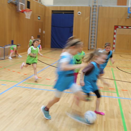 Eine Gruppe von Kindern in Sportkleidung spielt Fußball auf einem Hallenboden mit Holzbelag. Einige laufen, während andere den Ball jagen.