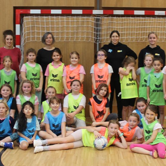 Eine Gruppe junger Mädchen in Sporttrikots posiert für ein Foto vor einem Tor mit erwachsenen Betreuern. Sie stehen auf einem Holzboden, und einige halten einen Fußball.