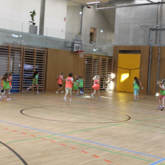 Eine Gruppe von Kindern in hellen Westen spielt ein Spiel auf einem Innenhof, mit Holzwänden und einer gelben Tür.