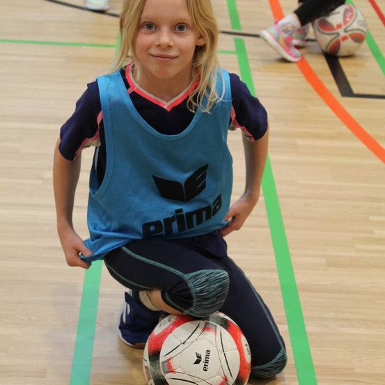 Ein Mädchen in einem blauen Trikot mit dem Wort 'prima' lächelt, während es auf einem Spielfeld kniet, mit einem Fußball vor sich.