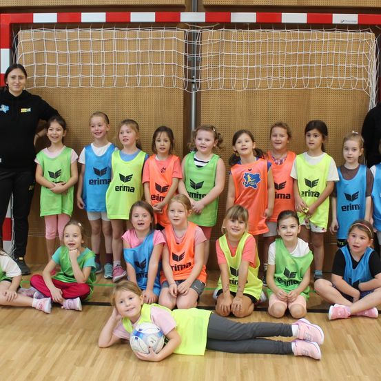 Eine Gruppe junger Mädchen in leuchtenden Trikots versammelt sich in einer Sporthalle. Einige stehen hinter einem Netz, andere sitzen oder knien auf dem Boden. Eine Frau steht lächelnd in der Nähe.