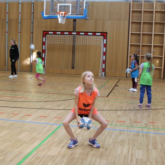 Eine Gruppe junger Mädchen spielt Volleyball in einer Turnhalle. Ein Mädchen in Orange hält einen Ball und schaut zur Seite. Ein Basketballkorb ist an der Wand montiert.