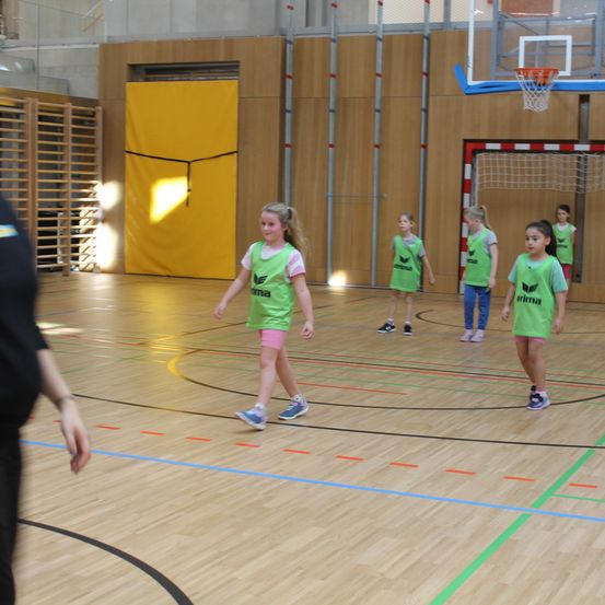 Eine Gruppe junger Mädchen in grünen Trikots spielt ein Spiel auf einem Hallenspielfeld. Dahinter befindet sich eine gelbe Tür.