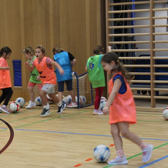 Eine Gruppe von Kindern spielt Fußball in Innenräumen, trägt leuchtend farbige Westen und Turnschuhe. Mehrere Bälle sind auf dem Spielfeld verstreut.